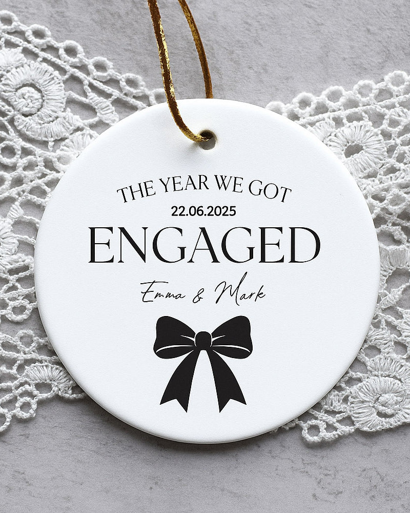Round ceramic ornament with 'The Year We Got Engaged' text and a bow design on a lace background.