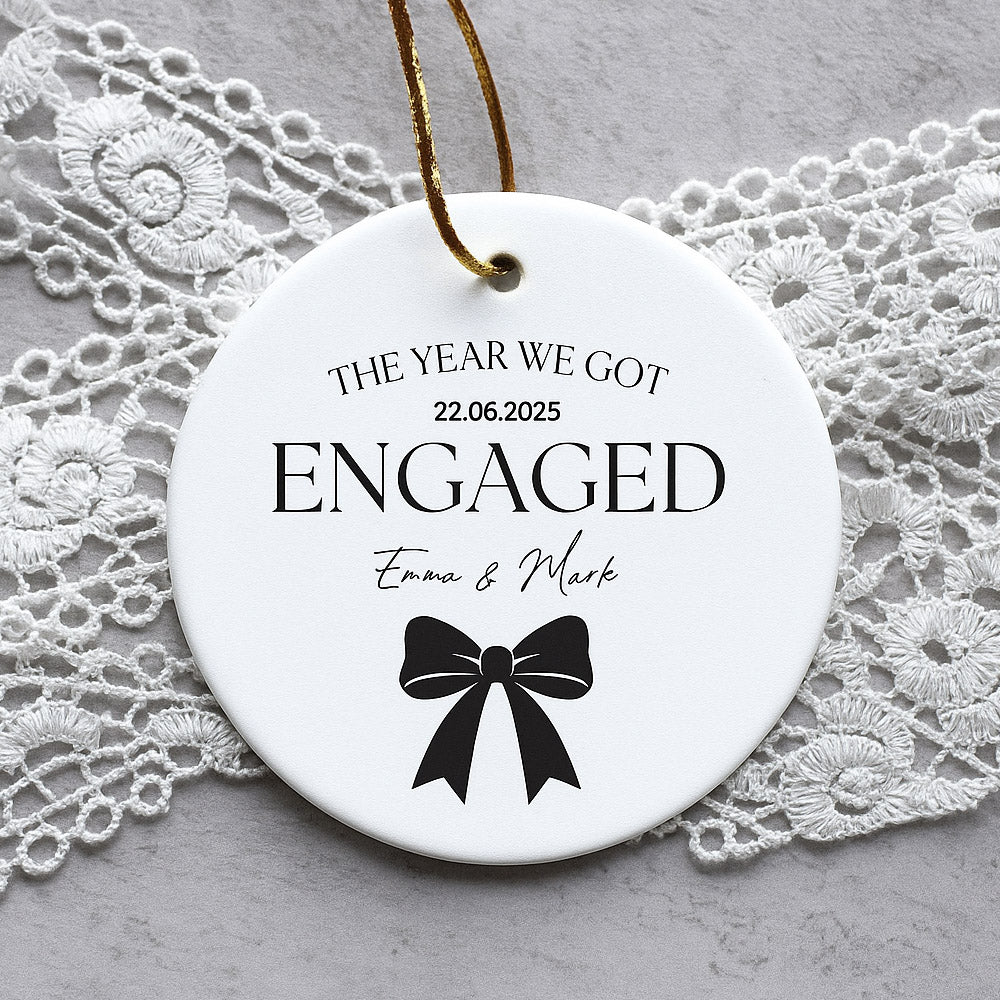 Round ceramic ornament with 'The Year We Got Engaged' text and a bow design on a lace background.