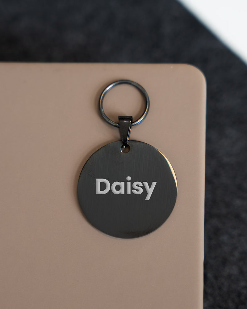 Black round pet tag with 'Daisy' engraved on it, attached to a beige surface.