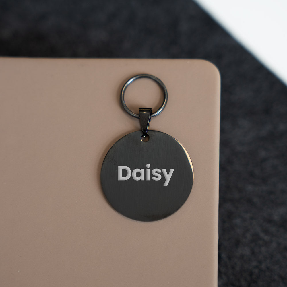 Black round pet tag with 'Daisy' engraved on it, attached to a beige surface.