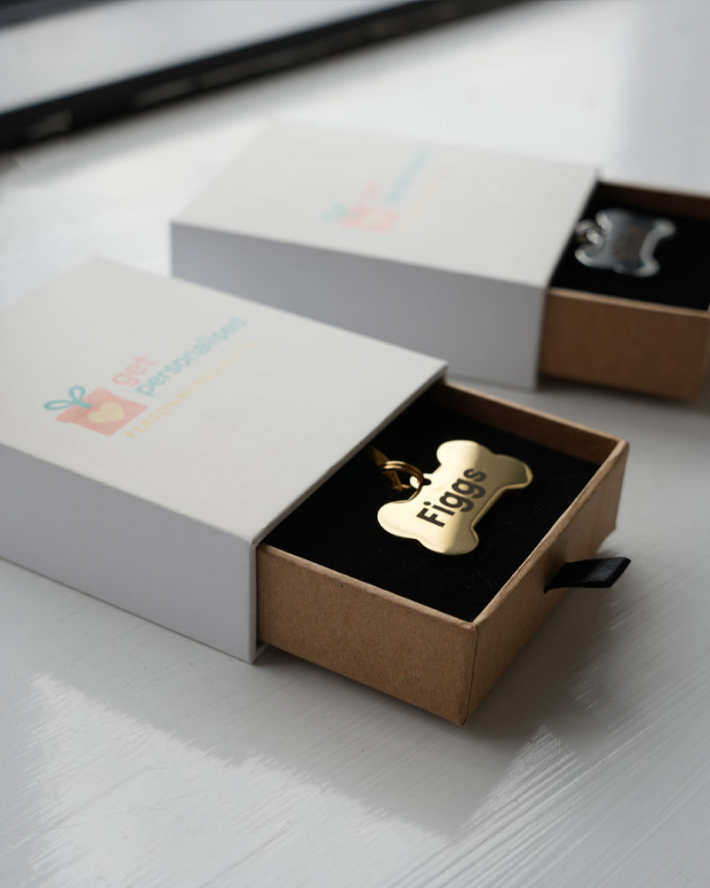 A gold metal pet ID tag in the shape of a bone, personalized with the name 'figgs' in luxury packaging.