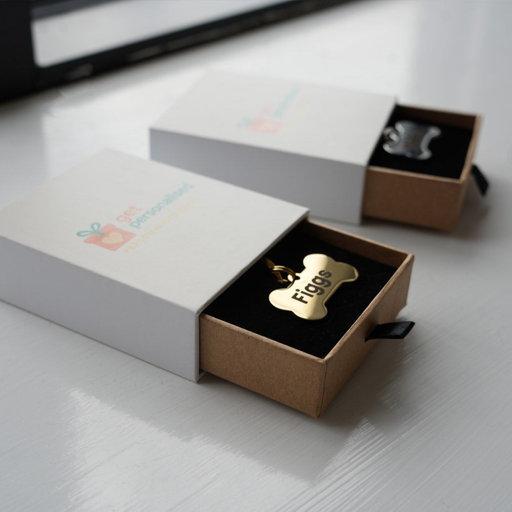 A gold metal pet ID tag in the shape of a bone, personalized with the name 'figgs' in luxury packaging.