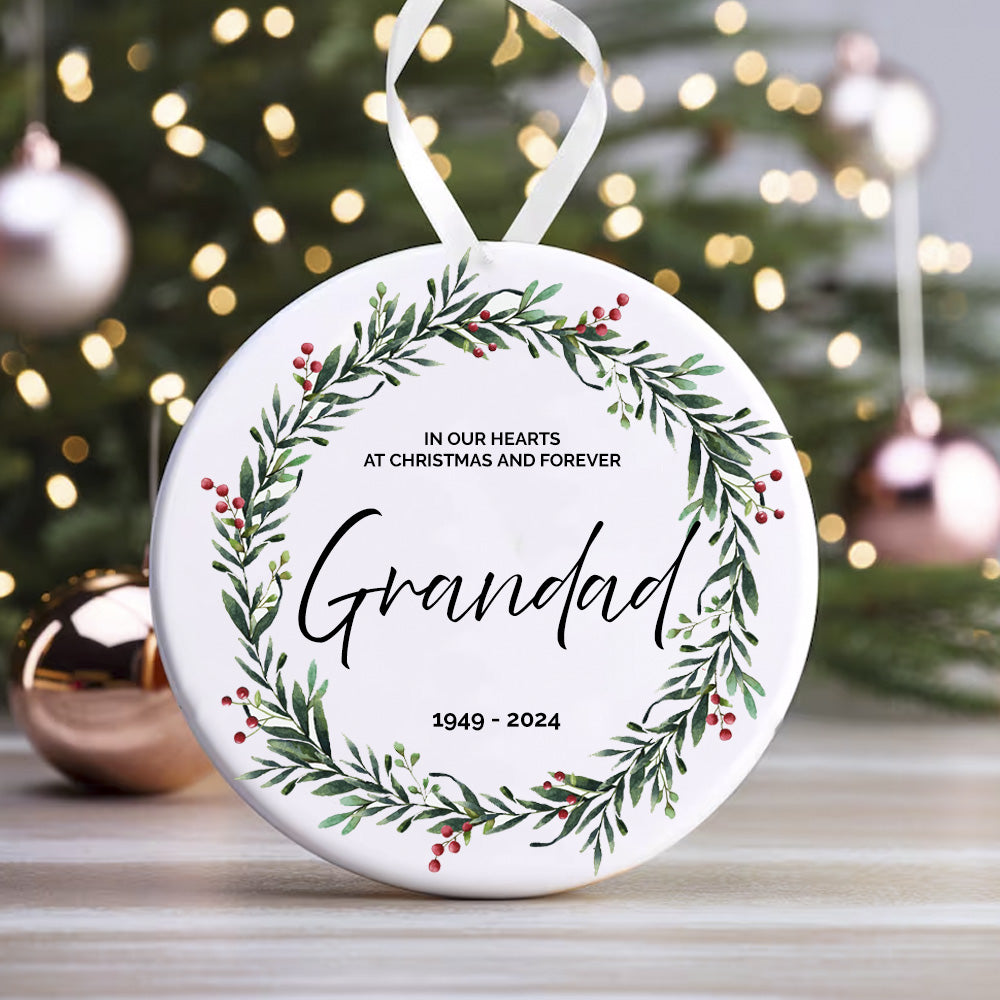 A round ceramic decoration with a wreath design and the text 'IN OUR HEARTS FOREVER AT CHRISTMAS AND FOREVER' along with a space for personalization, hanging against a blurred Christmas tree and holly decoration background.