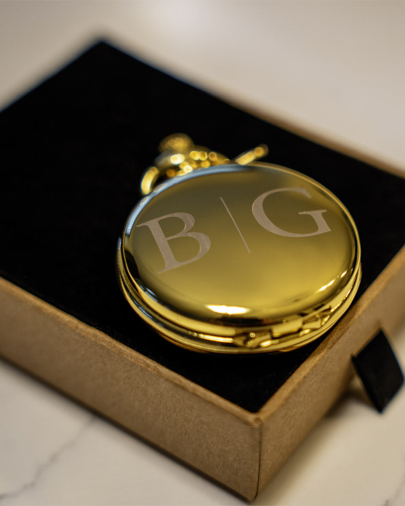 Gold pocket watch with 'B&G' initials in a box on a marble surface