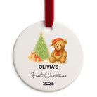 Round Christmas ornament with a teddy bear, tree, and presents, personalized for 'Olivia's First Christmas 2025'.